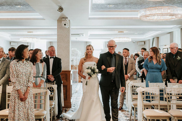 a bride walking down aisle on arm of man in black suit during wedding ceremony at Busby Hotel