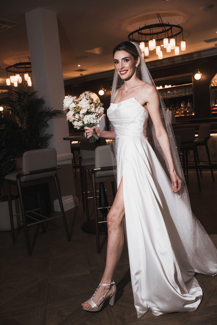 a smiling bride holding a rose bouquet walking confidently through a softly illuminated dark room.