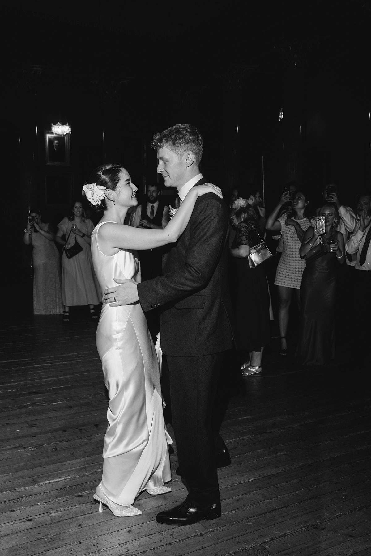 Black and white photo of bride and groom sharing their first dance surrounded by guests.