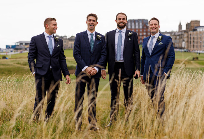 Four smiling men in suits with boutonnieres standing in a field of tall grass with the Old Course Hotel in the background