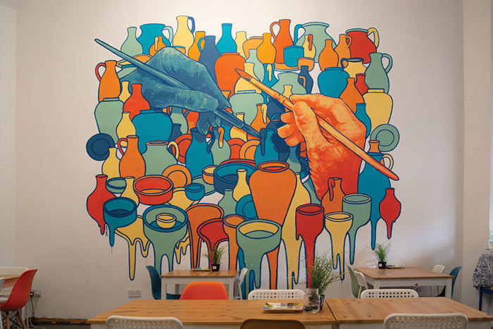 a bold coloured wall mural depicts two large hands each holding a paint brush and lots of ceramic pots and vases around them with the bottom row looking to be dripping paint