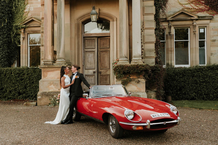 Bride and groom lean into each other posing in front of red classic Jaguar convertible