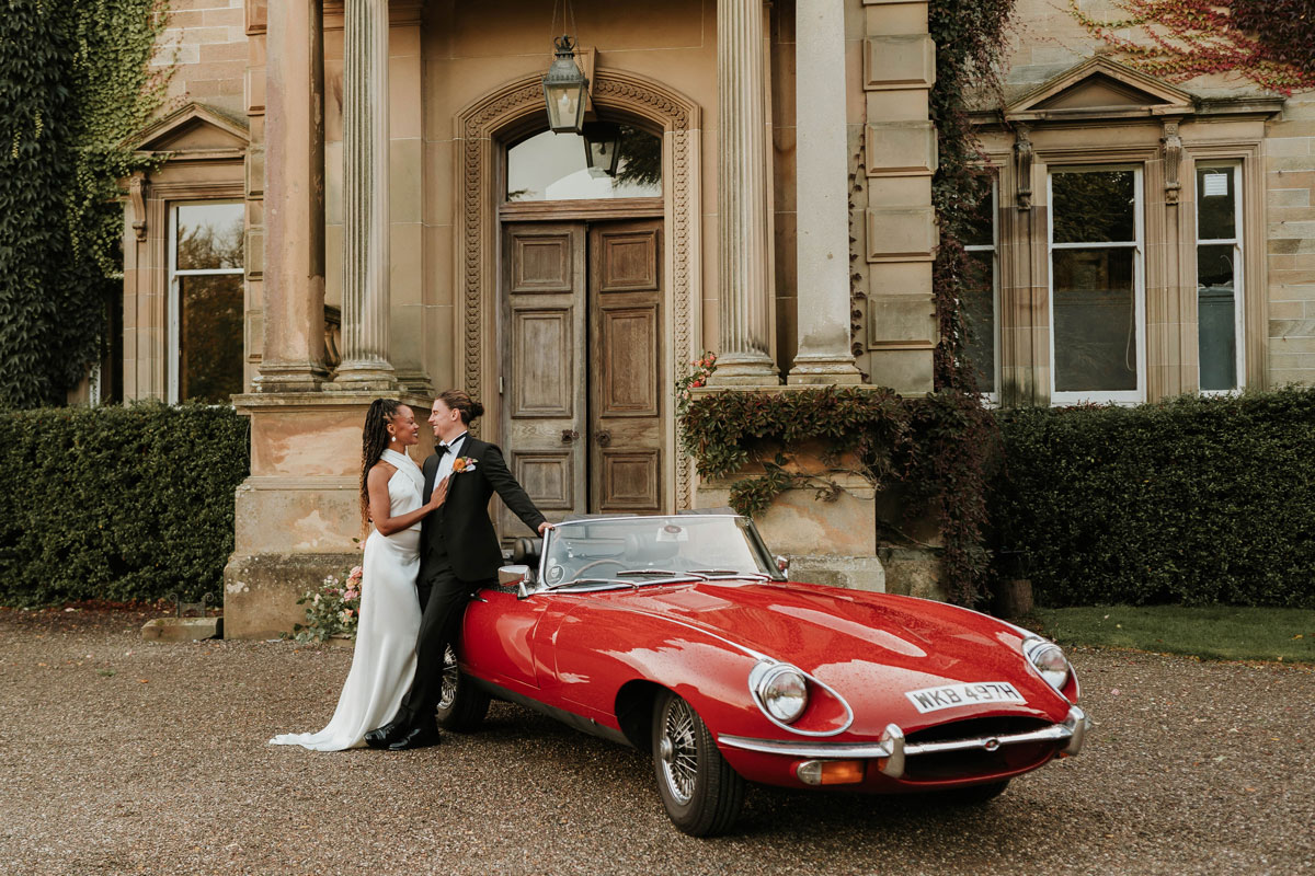 Bride and groom lean into each other posing in front of red classic Jaguar convertible
