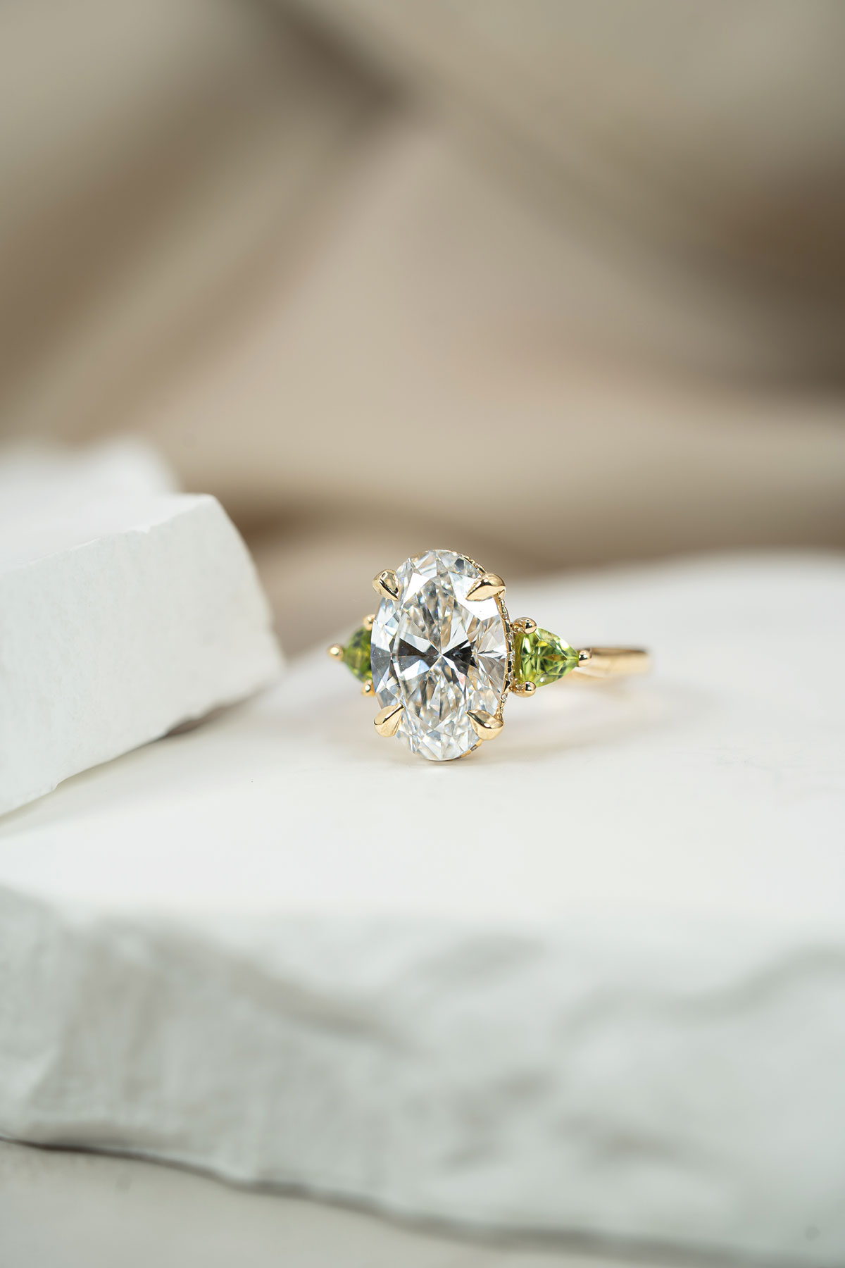 Bespoke oval-cut engagement ring with green gemstone side stones on a gold band by Paragon Diamonds