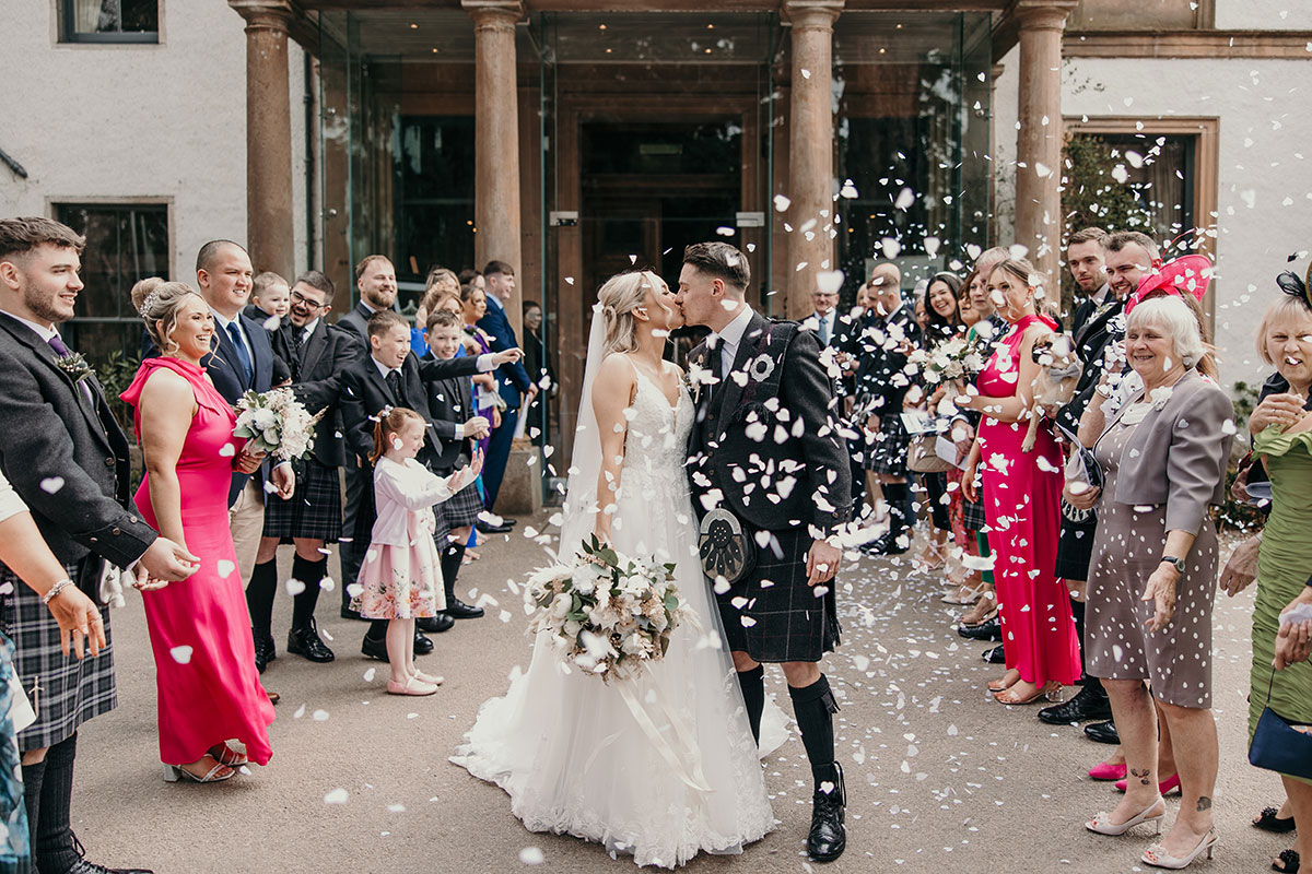 bride in thin strapped flowy wedding dress and holding neutral bouquet kisses groom in full kilt outfit surrounded by white flower petals floating in the air with a row of friends and family stood on either side of them smiling after having thrown the petals