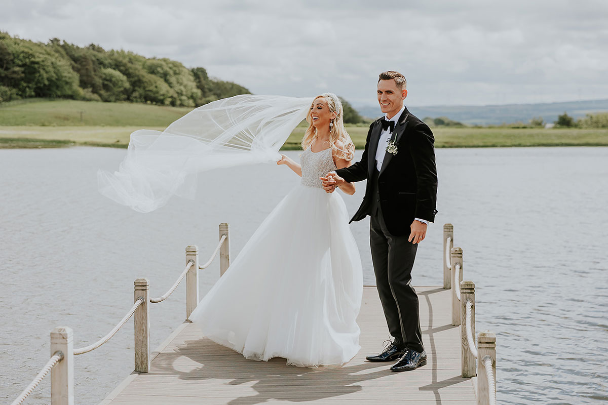 blonde bride in ballgown with windblown veil and groom in tux hold hands on a dock with calm waters around them