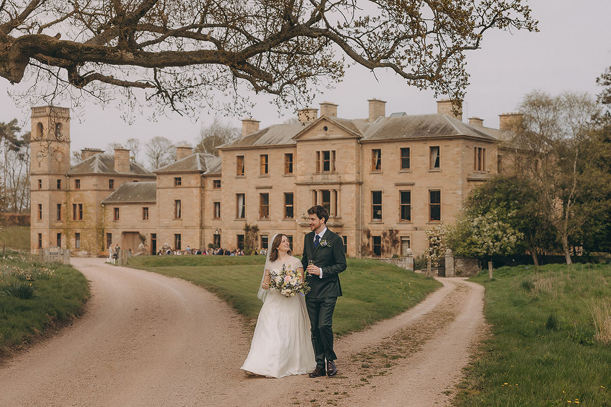 A bride and groom walking along a curved driveway in front of a large historic house with guests gathered outside.