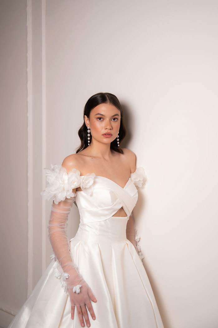 bride with dark brown wavy hair wears strapless off-the-shoulder wedding dress with cut out bodice detailing and long floral applique mesh sleeves as she stands in front of a white wall