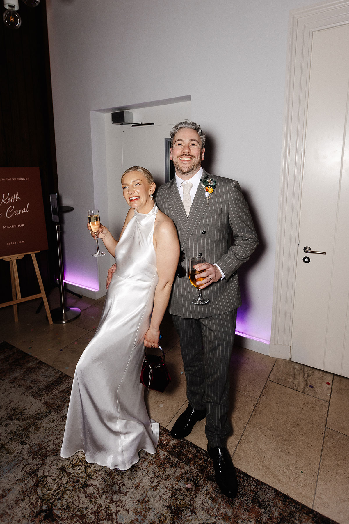 Bride in a satin halterneck dress and groom in a grey pinstripe suit at Virgin Hotels Edinburgh, smiling during their modern city wedding reception.