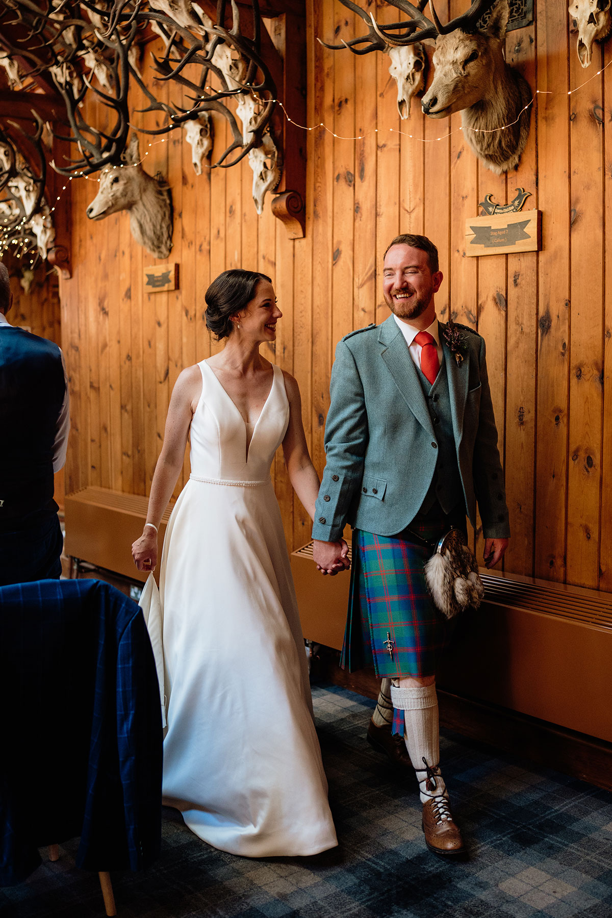 Bride and groom walking hand in hand through the Great Hall at Mar Lodge Estate beneath its famous antler-lined ceiling