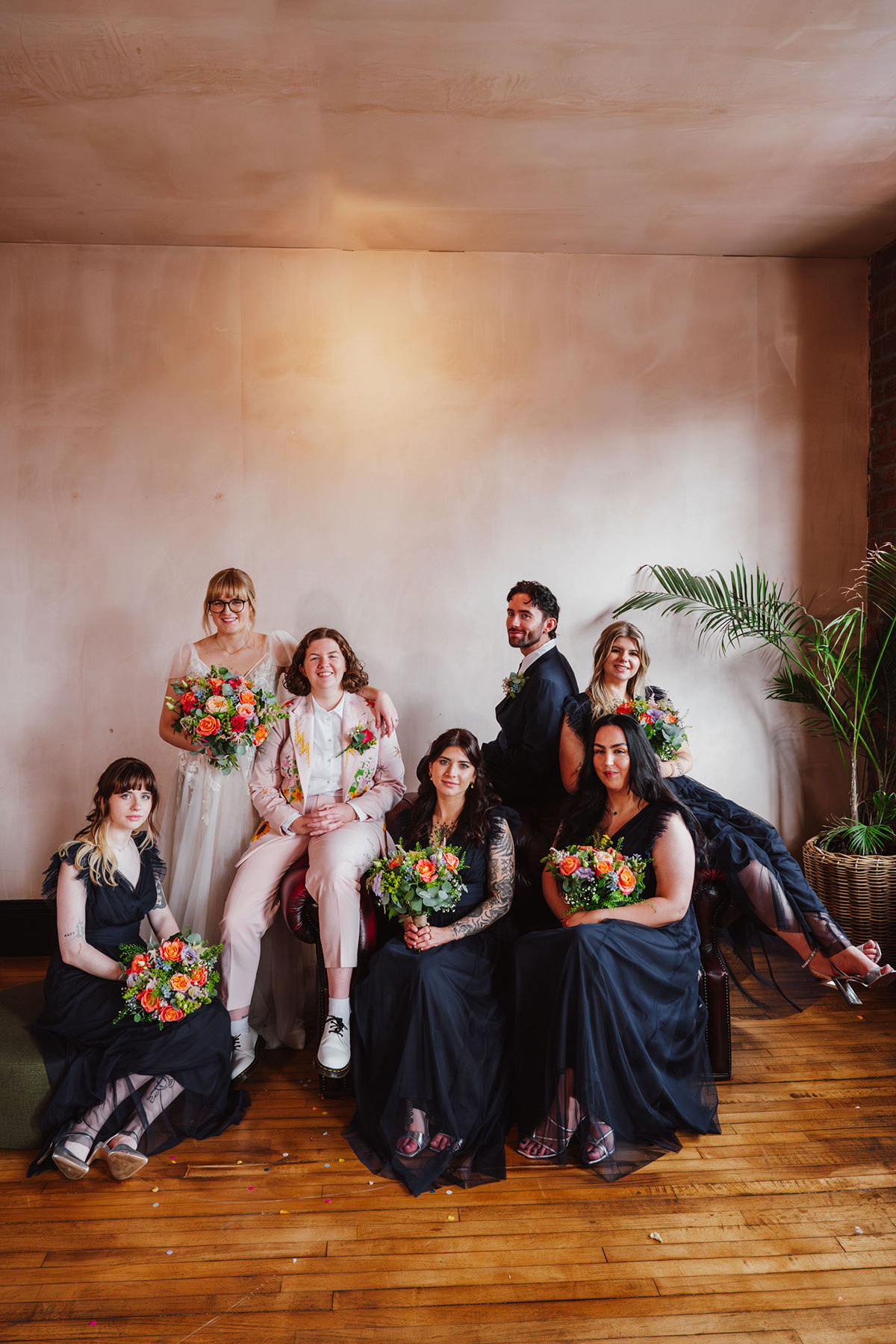 Bridal party group portrait inside the Engine Works, with both brides and their attendants in coordinating outfits holding colourful bouquets.