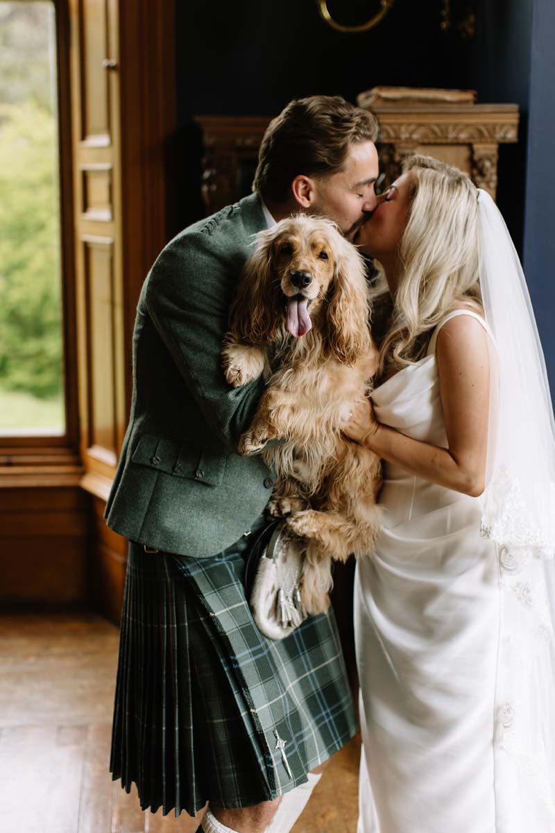 Newlyweds Lia and Louis kiss while holding their cocker spaniel Bertie at their Cambo House wedding.