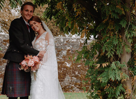 bride and groom both smile as they stand outside surrounded by autumnal foliage and lean into each other, the bride resting her head on the groom's lower shoulder