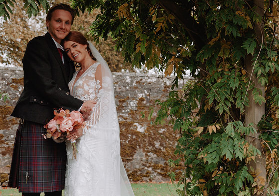 bride and groom both smile as they stand outside surrounded by autumnal foliage and lean into each other, the bride resting her head on the groom's lower shoulder