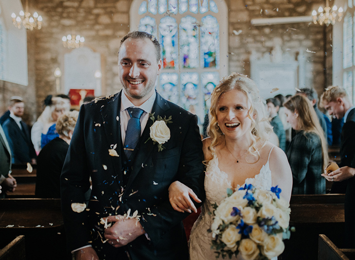 A bride and groom walking arm in arm out of a church as confetti is being thrown on them