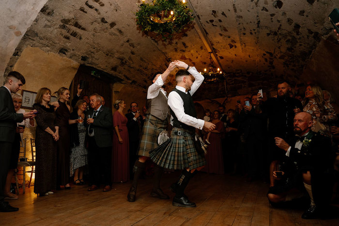 two grooms in kilts doing a first dance in a room at Neidpath Castle as guests watch on