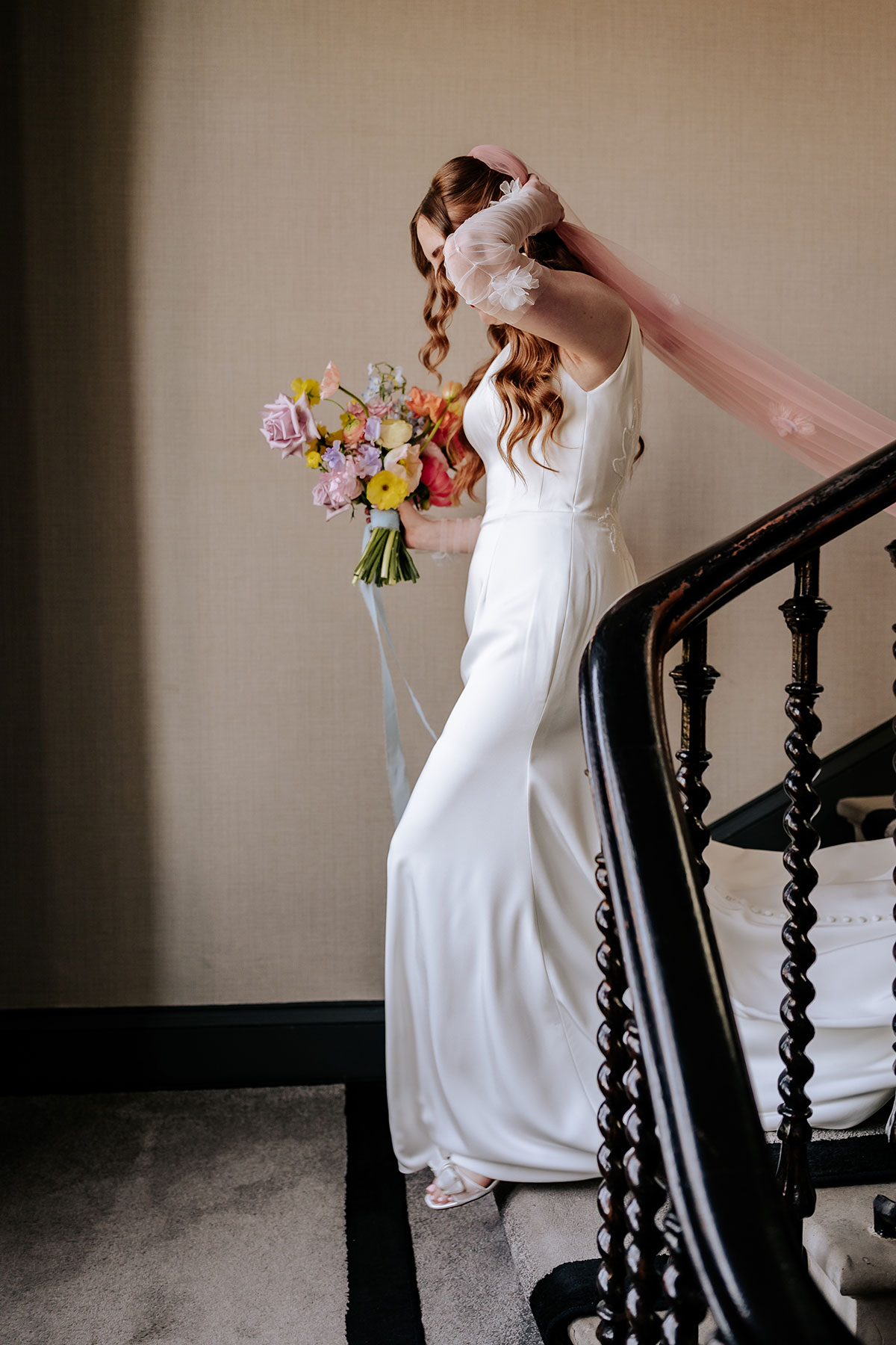a bride wearing a pink veil and carrying a colourful bouquet of flowers walking down a set of stairs
