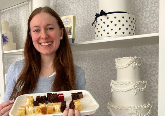 a brown haired woman stands in front of shelves with wedding cakes holding a tray of tiny different flavoured cake slices