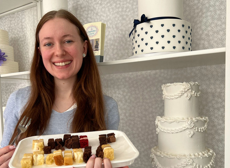 a brown haired woman stands in front of shelves with wedding cakes holding a tray of tiny different flavoured cake slices