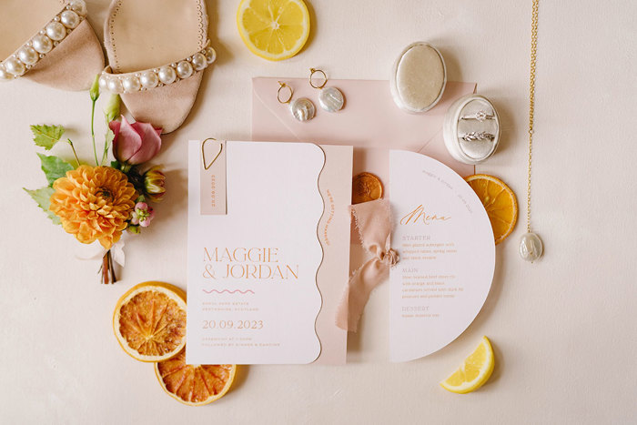 a wedding flatlay including pink and white stationary, pearl heels and jewellery and flowers plus citrus