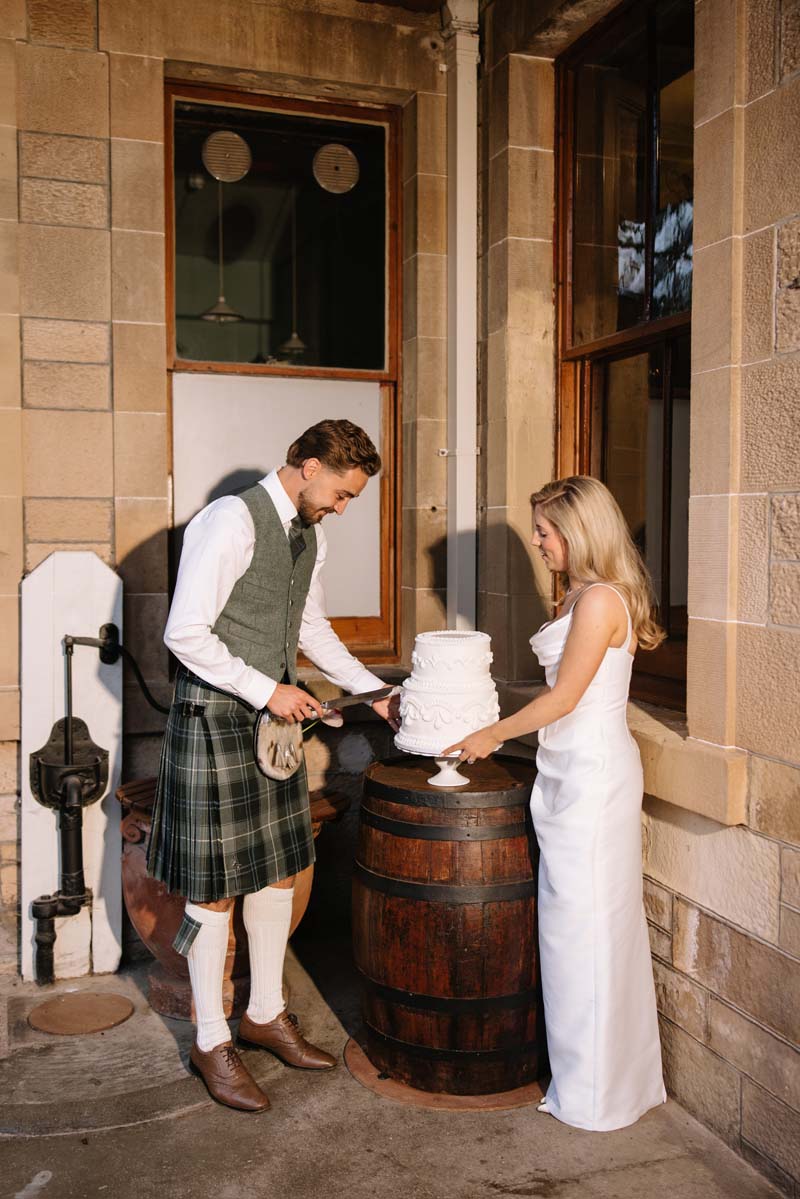 Lia and Louis cut their white two-tier wedding cake outside Cambo House during their wedding reception.