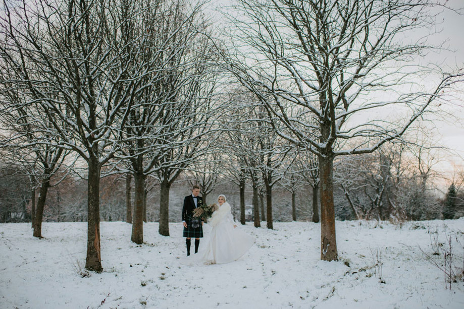 A bride wearing a white dress and white fur wrap holding hands with a groom wearing a dark kilt walking in a park in the snow