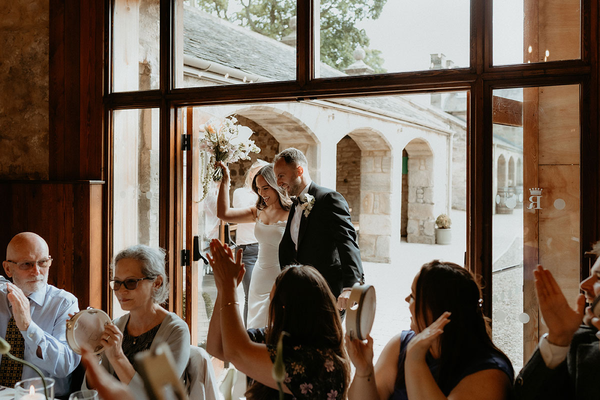 Bride and groom entering wedding breakfast at Rosebery Steading, Midlothian, greeted by guests with tambourines.