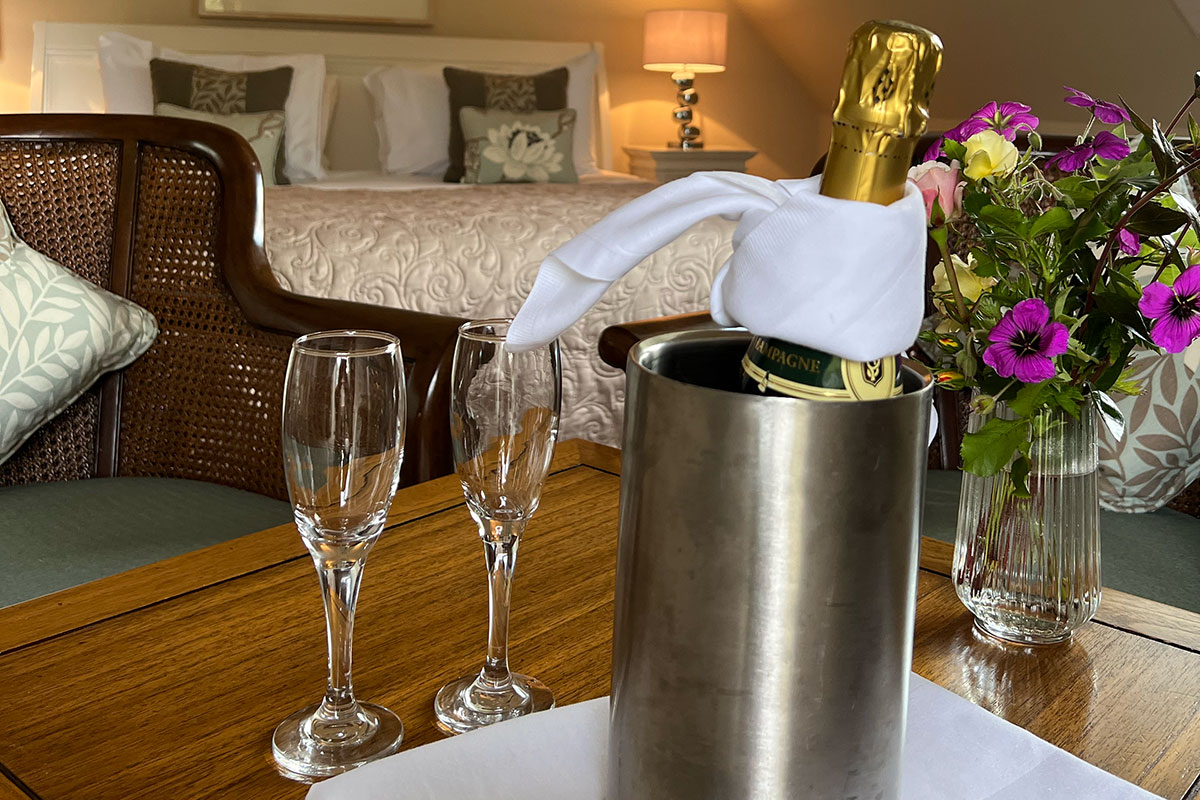a chilled bottle of champagne and two empty glasses sit on a wooden table with a large well-made bed in the background