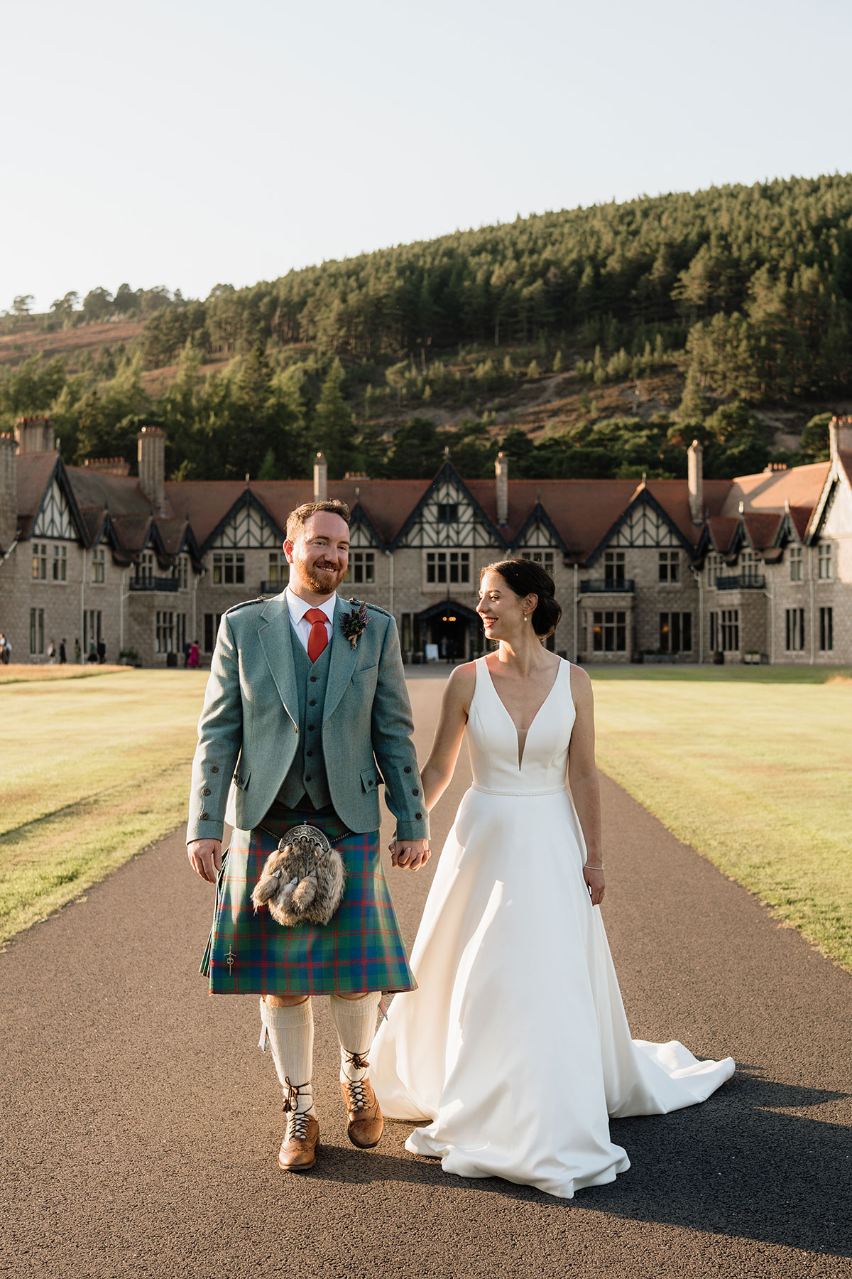 Bride and groom walking together outside Mar Lodge Estate with the historic Highland lodge and Cairngorm scenery behind them