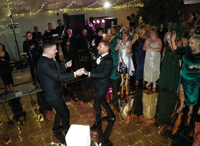 two grooms dancing on a gold mirrored dance floor as gathered guests stand on the side clapping. There is a wedding band on the stage behind them