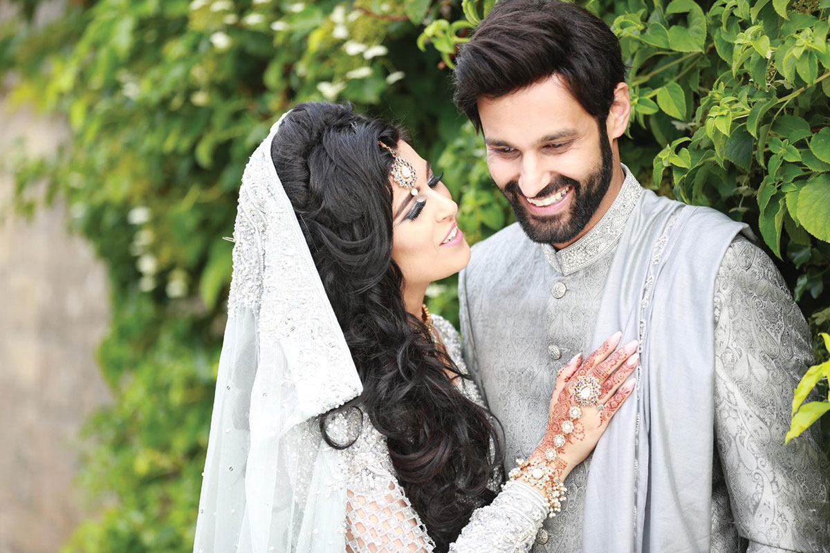 Bride and groom in traditional South Asian wedding attire standing close together in a garden; the bride wears an embellished outfit with veil and intricate henna, and the groom wears a silver sherwani