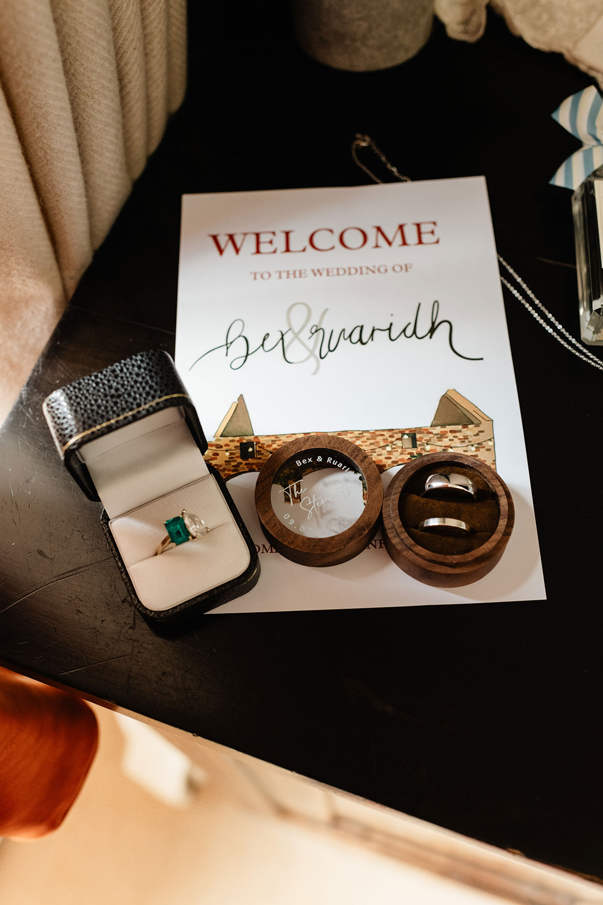 Wedding rings displayed in wooden ring boxes beside an emerald-cut engagement ring and personalised wedding welcome card for Bex and Ruaridh