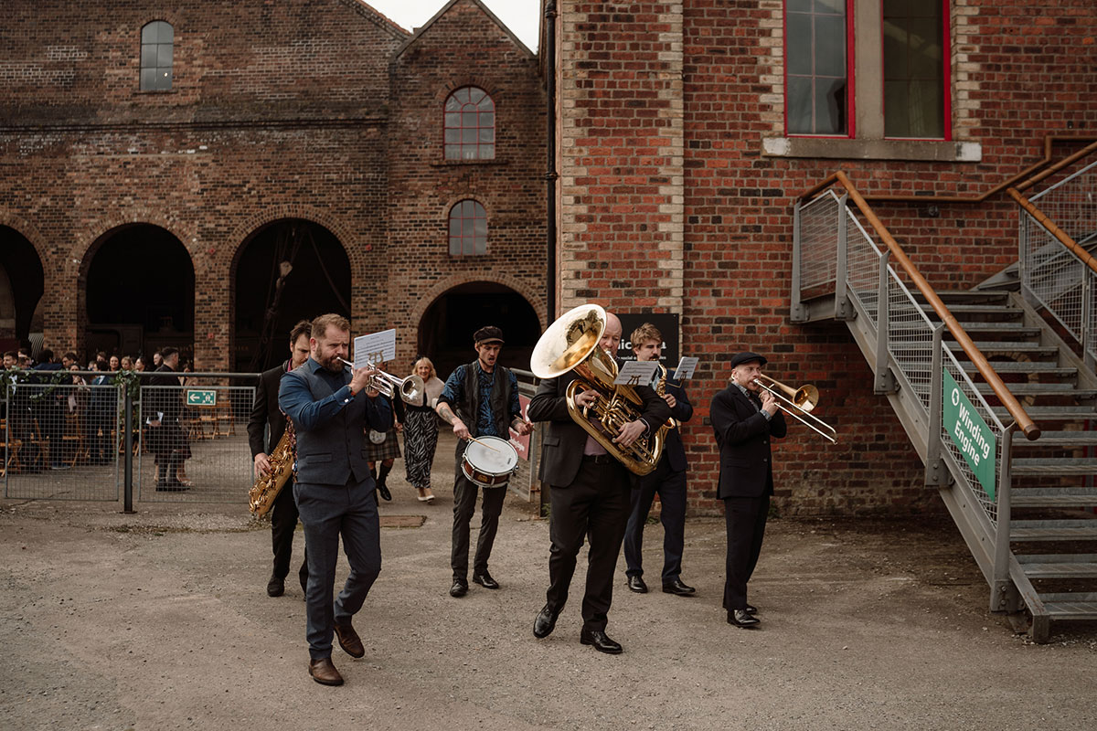 Brass band performs outdoors during a wedding celebration at a historic industrial venue, entertaining guests between ceremony and reception