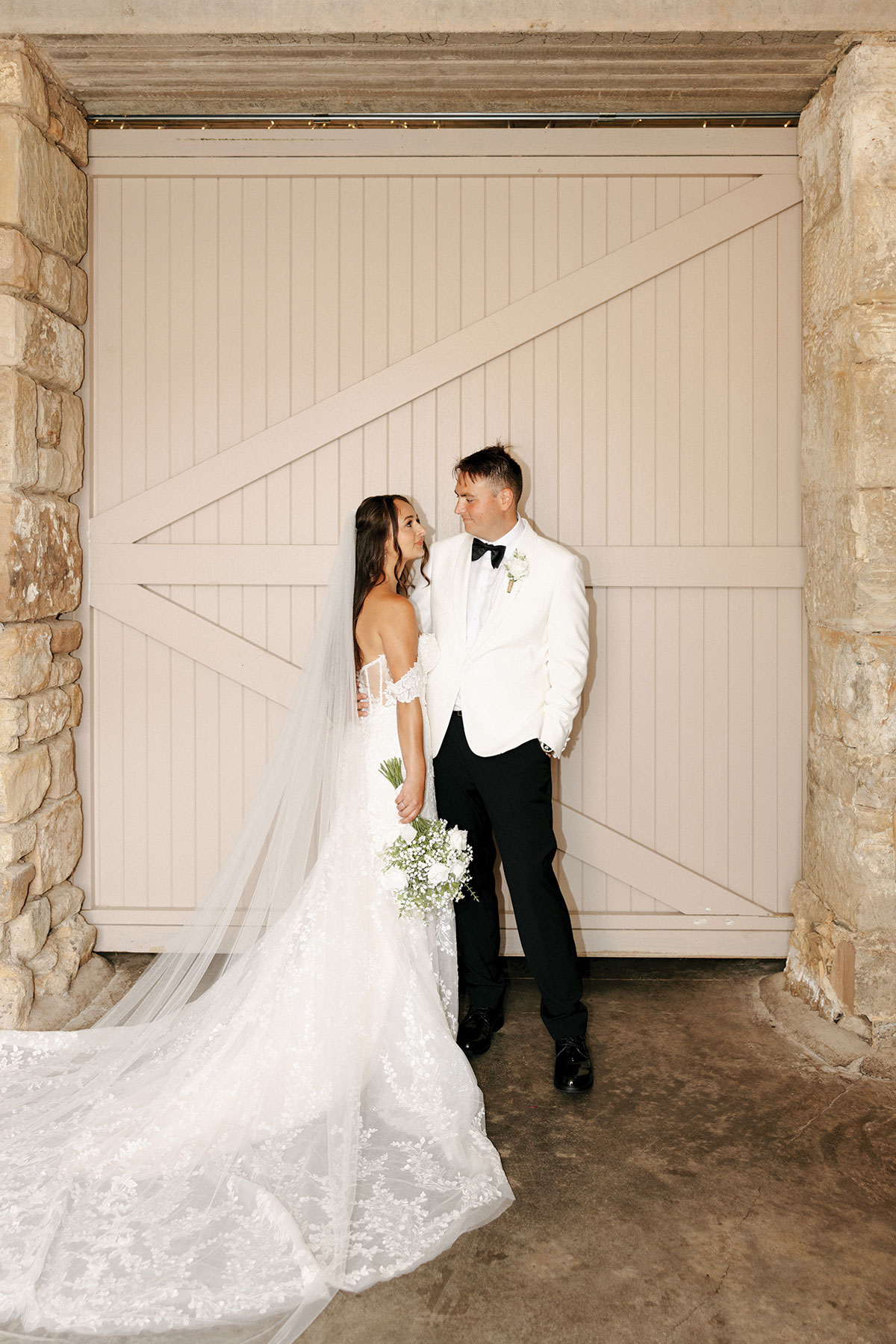 Bride and groom pose together at Falside Mill, standing by a light-coloured barn door framed by stone pillars.