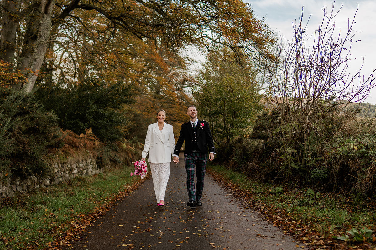 Bride and groom walk hand in hand along autumn country road during Scottish wedding photoshoot