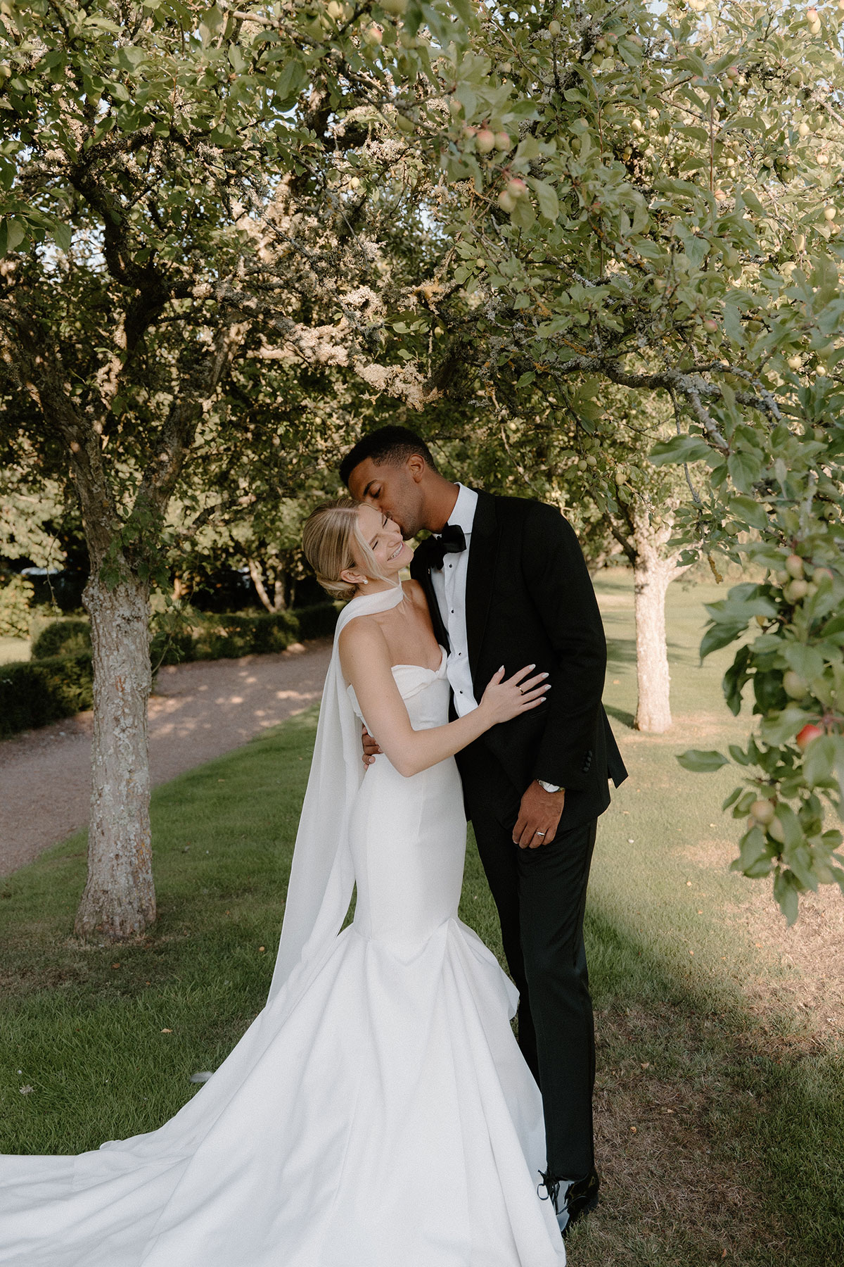 Bride and groom posing beneath apple trees in the gardens of a Scottish wedding venue, with the bride wearing a fitted satin gown and scarf detail