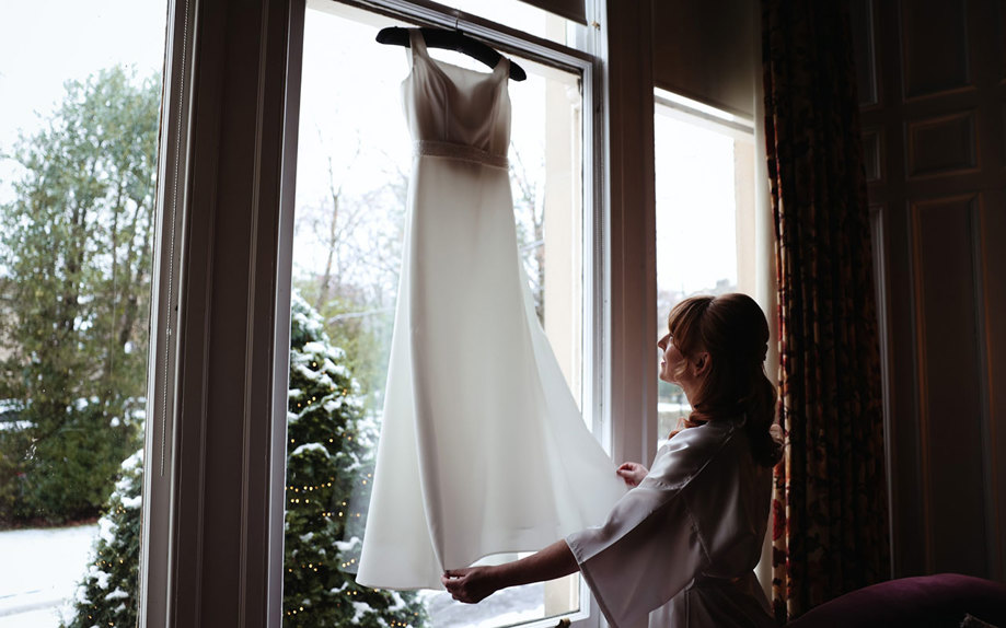 a wedding dress hanging up in front of a window with a snowy Christmas tree outside and a woman in a white robe holding the edges of the dress