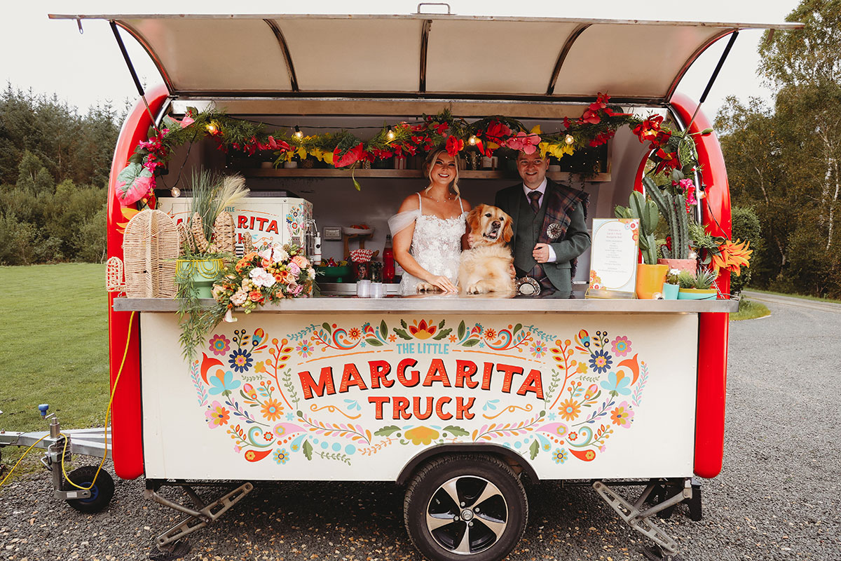 Bride, groom and their golden retriever posing behind a colourful margarita truck at a Scottish wedding venue