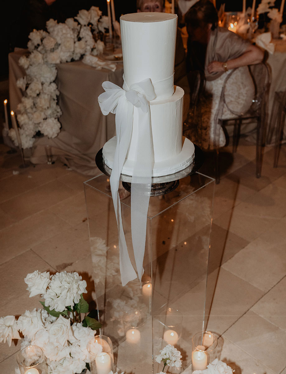 a two-tier white wedding cake with large bow tied around it sitting on a clear perspex stand surrounded by white flowers and soft candlelight