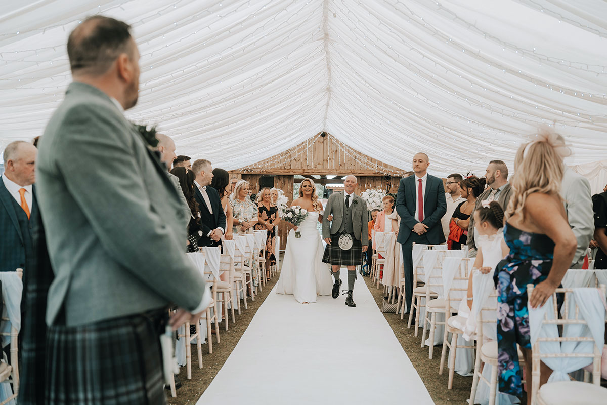 Bride walking down the aisle of a marquee ceremony with her father as guests look on.