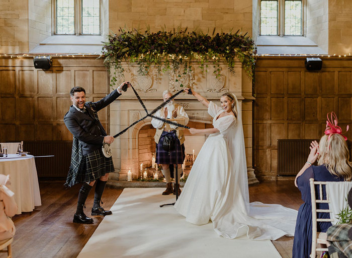 a bride and groom look elated and hold tartan ribbons aloft as they stand in front of a large engraved stone fireplace with officiant clapping