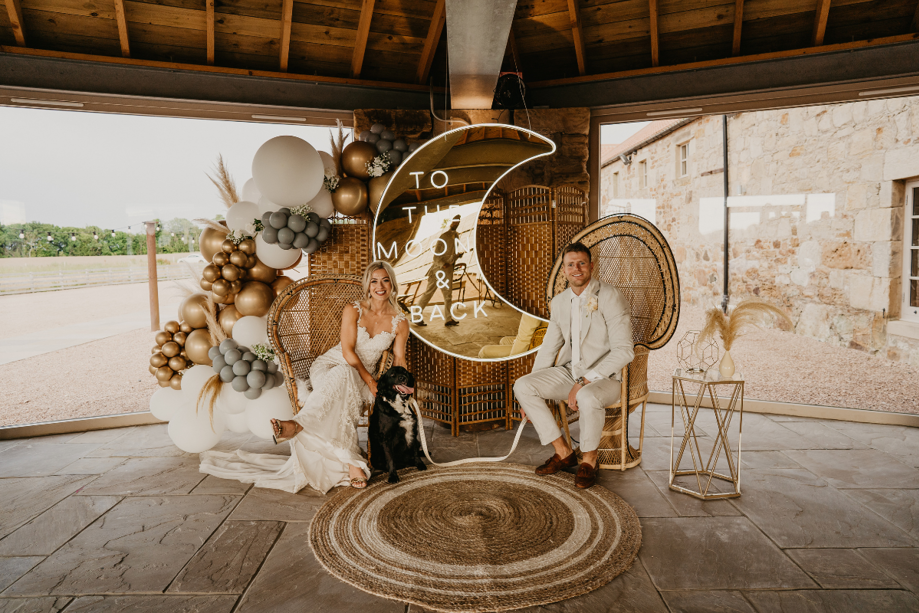 Man and woman sitting o rattan chairs with dog and sign that says 'To the moon and back'