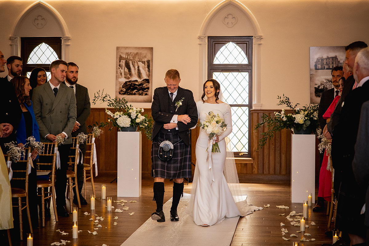 Bride smiles as her father walks her into the ceremony room filled with guests