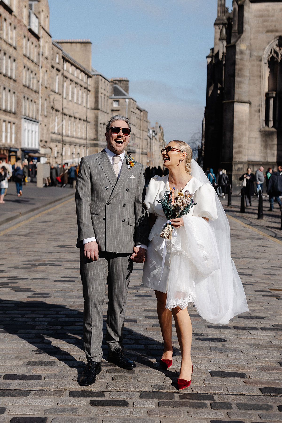 Newlyweds laugh together on Edinburgh’s Royal Mile, the bride in a short puff-sleeve dress and red heels, the groom in a grey pinstripe suit.