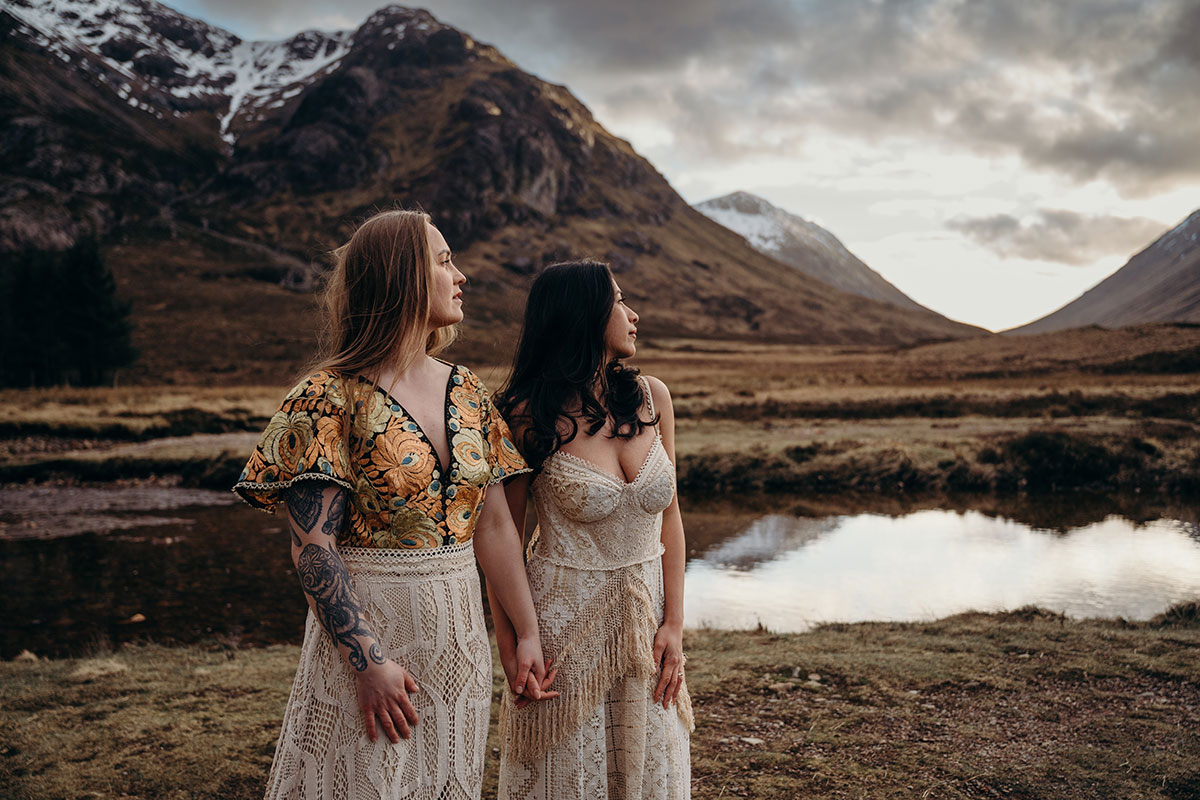 Couple holding hands beside reflective water during a Glencoe elopement shoot with Scottish mountain scenery.