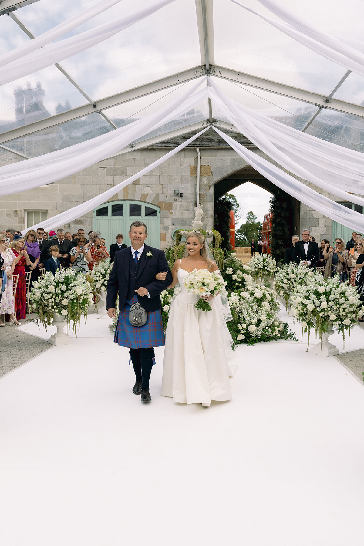 Bride walks down the aisle with her father beneath a draped canopy and floral arch at Dundas Castle courtyard.
