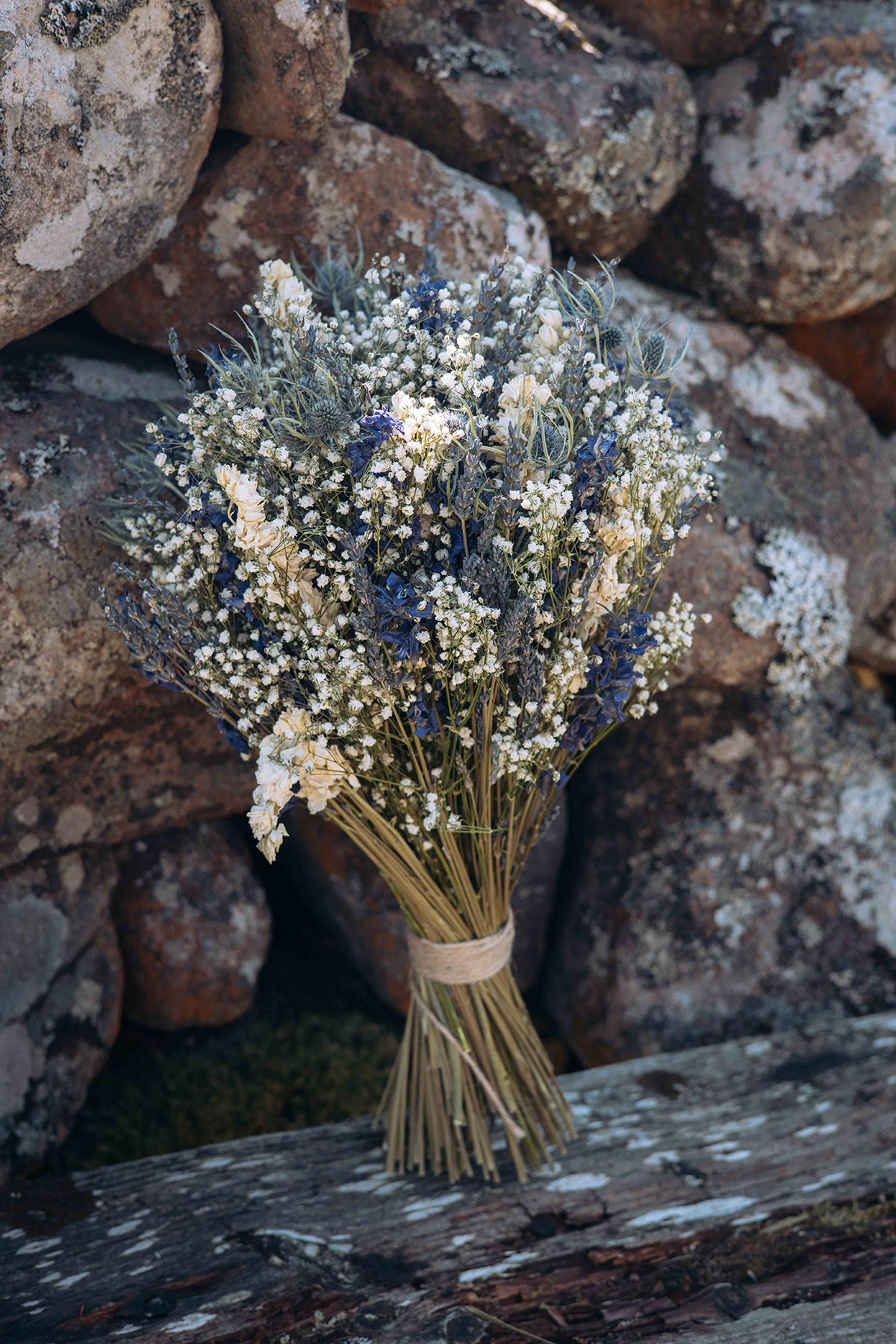 Rustic dried wedding bouquet of lavender, thistle and baby’s breath, tied with twine against a stone wall in the Scottish Highlands.