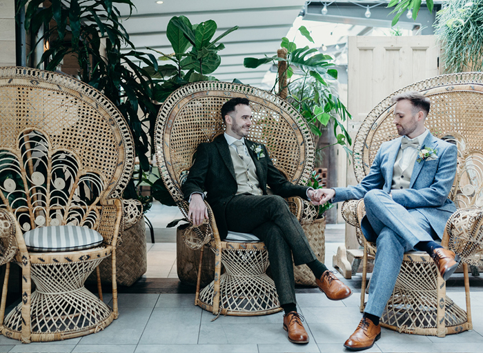 two grooms holding hands sitting with their legs crossed on wicker peacock chairs with plants in background