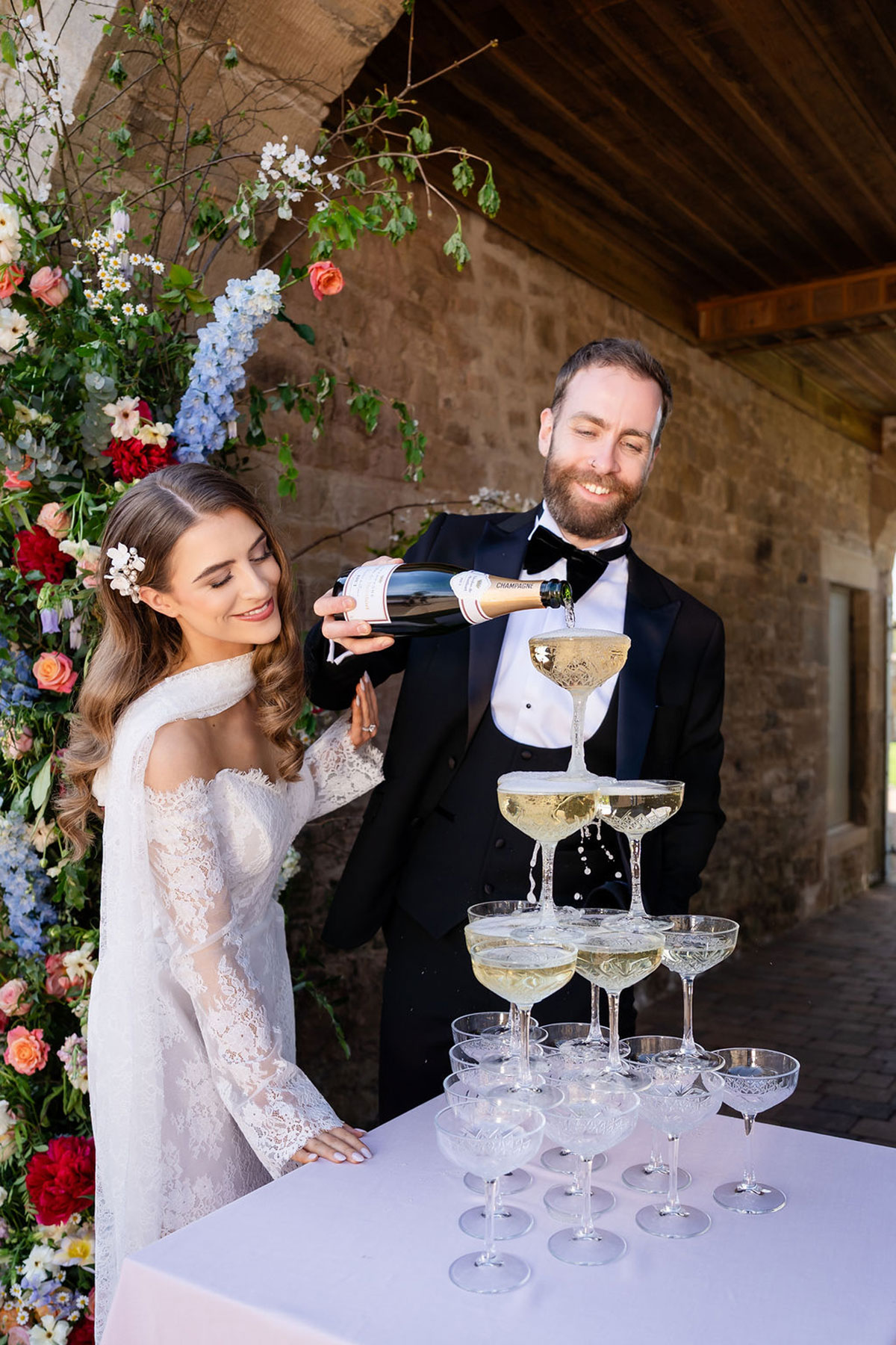 Bride smiling as the groom pours champagne into a coupe tower, surrounded by colourful florals against a rustic stone backdrop