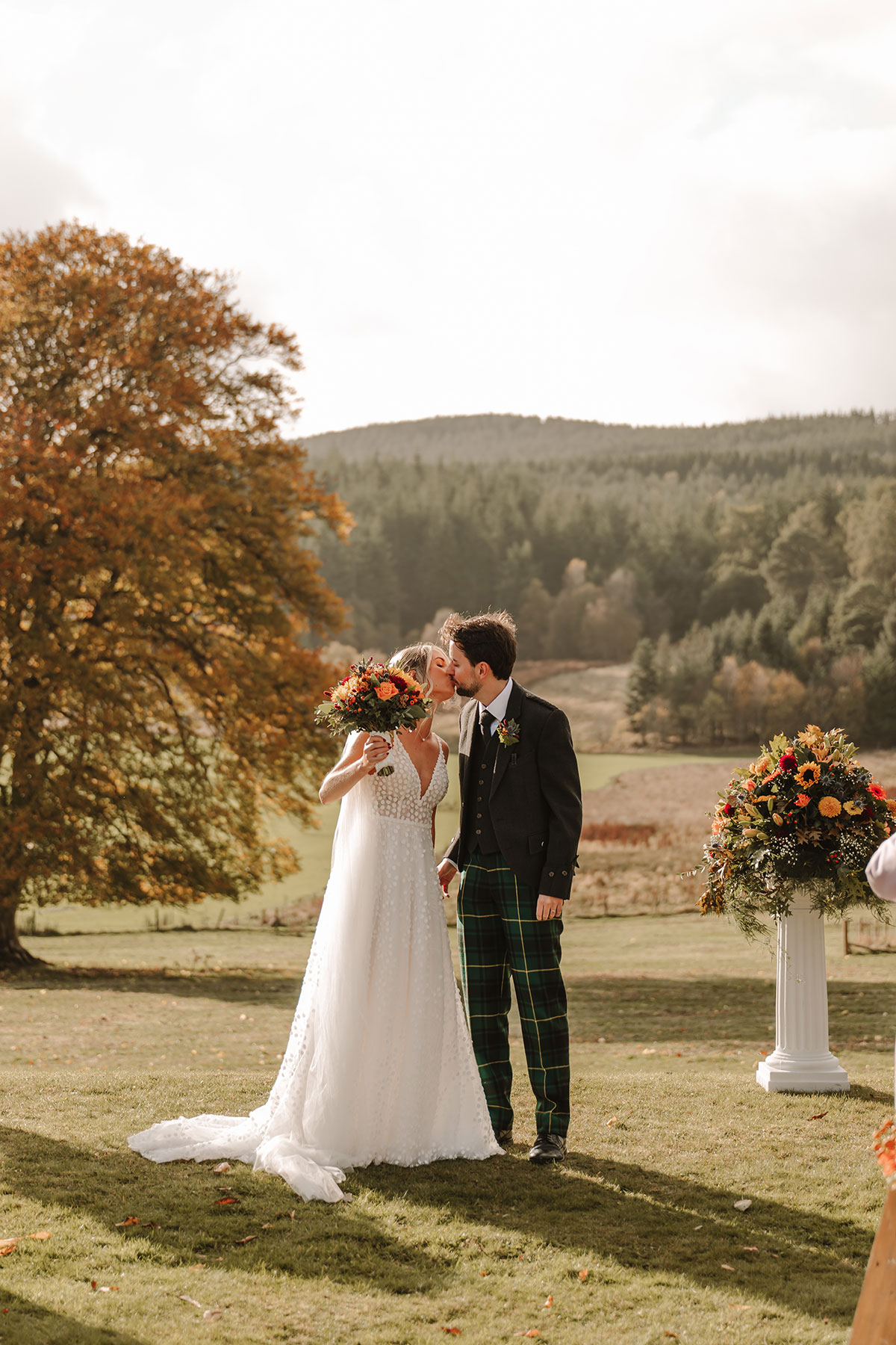Bride and groom kissing outdoors after wedding ceremony with autumn trees in the background.